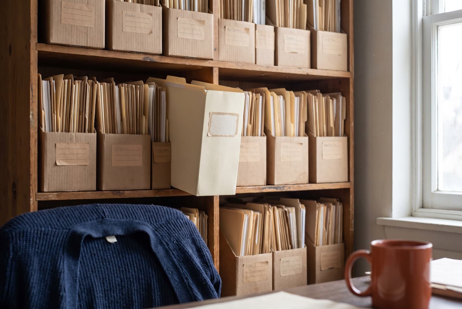 Labelled file boxes and manila folders on an open wooden shelf, one pulled forward.