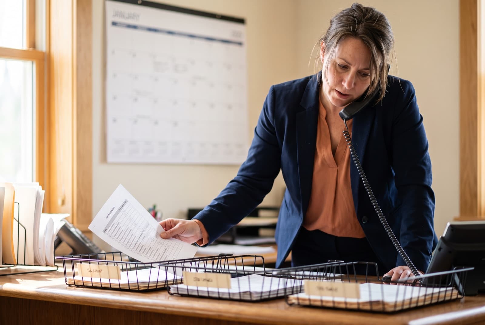 A manager on the phone at a desk holding a printed checklist, three document trays in front of her.